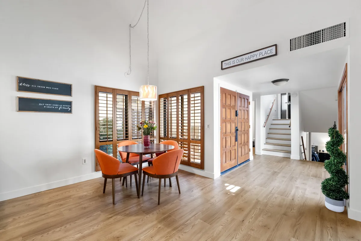 A dining area with a round table and vibrant orange chairs, situated near the entrance door and complemented by decorative wall signs. The setup is ideal for meals, casual conversations, and fostering a sense of community and connection among guests.