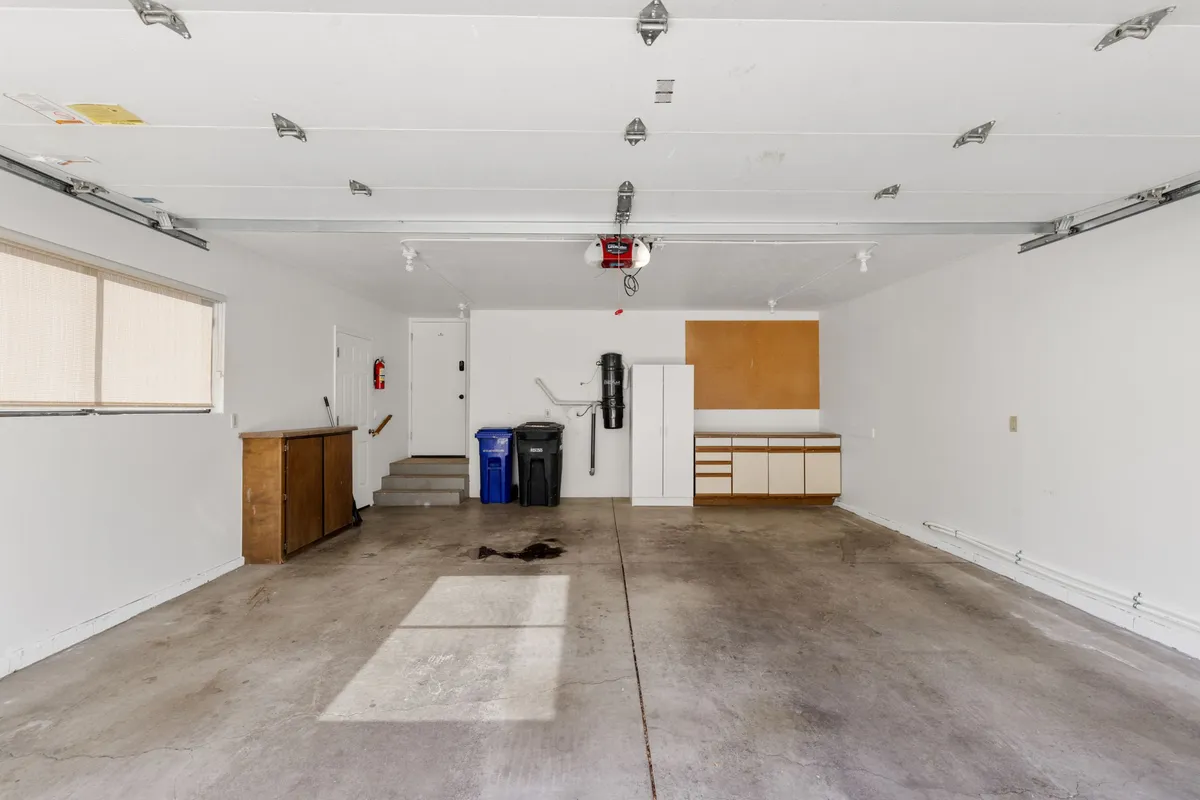 An organized garage space with cabinets, a garage door, windows, and stairs, offering ample room for storing bikes, hiking gear, and outdoor equipment. The concrete floor and visible storage solutions make it clear this area is practical for preparing and launching outdoor adventures.