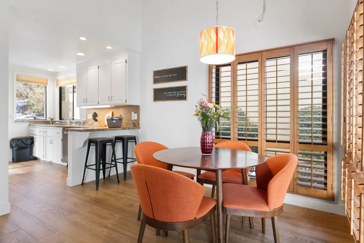 A warm dining space with a round wooden table surrounded by orange chairs, a vase of fresh flowers, and a pendant light overhead. Sunlight pours in through a shuttered window, infusing the room with a cozy, inviting glow perfect for shared meals and morning coffee.