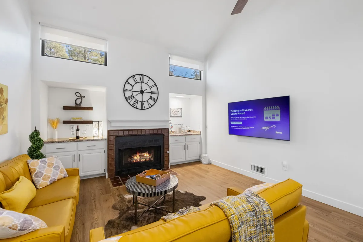 A bright, cheerful living area featuring a yellow sofa, a fireplace, a wall clock, and a TV screen. The space is designed for gatherings, with comfortable seating and a warm, inviting atmosphere ideal for relaxing, entertaining, or movie nights.