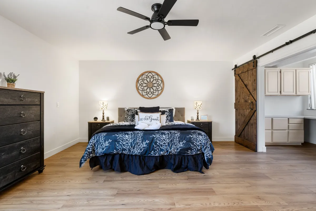 A calm and welcoming bedroom with blue floral bedding, a wooden dresser, and a rustic barn door. The space exudes a peaceful, homey feel, perfect for quiet moments, morning routines, or winding down.